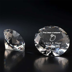 Puede incluir: Dos adornos transparentes de cristal con forma de diamante. Uno es parcialmente visible, el otro está a la vista, con el texto "Nos âmes s'unissent" y "Clara & Hugo Fiançailles 2024" grabado, junto con un gráfico de anillo.