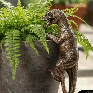 May include: A bronze-effect dinosaur ornament clinging to a dark grey plant pot overflowing with green ferns. The sculpture has intricate detailing and appears to be interacting with the plant. The pot is made of a dark, textured material, creating a naturalistic scene.