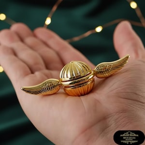 May include: A golden Snitch replica with detailed wings, a spherical body, and a ribbed design. The collectible is a shiny gold color and is a replica of the Quidditch ball from the Harry Potter series.