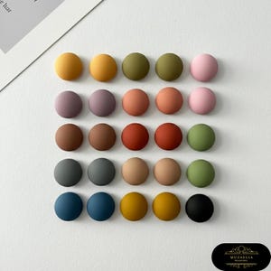 May include: A selection of twenty-four round, matte-finish decorative buttons in a variety of colours, including yellow, green, pink, and blue. The buttons are arranged in a grid pattern, offering a palette of hues for craft projects.