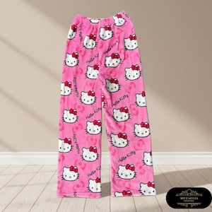 May include: Pink pajama pants featuring a repeating pattern of Hello Kitty's face with a red bow. The words "Hello Kitty" are printed throughout the design. The pants are made of a soft, plush material.