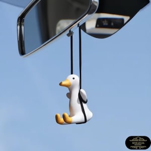 Könnte beinhalten: Ein weißer Enten-Autoanhänger mit gelben Füßen und Schnabel, der an schwarzen Schnüren hängt. Die Ente hängt vor dem Rückspiegel eines Autos vor blauem Himmel. Der Anhänger verleiht jedem Fahrzeug einen Hauch von Laune.