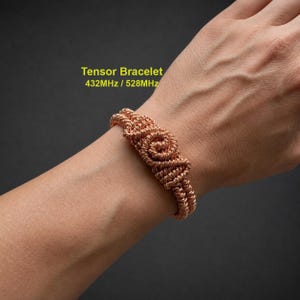 May include: A copper-colored wire bracelet featuring a central coiled design. The text "Tensor Bracelet 432MHz / 528MHz" is printed in yellow above the bracelet. The bracelet is constructed from multiple strands of twisted metal wire.