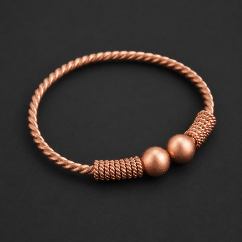 May include: A copper-colored bangle bracelet featuring a twisted rope-like texture throughout the band. The bracelet is accented with two smooth, spherical beads positioned between two sections of coiled wire detailing on one side of the circular frame.