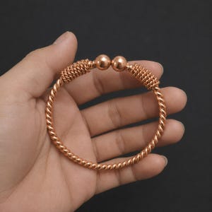 May include: A copper-colored metal cuff bracelet featuring a twisted rope design throughout the band. The open-ended bracelet terminates in two smooth, polished spherical beads, with a coiled wire texture accenting the ends near the spheres.