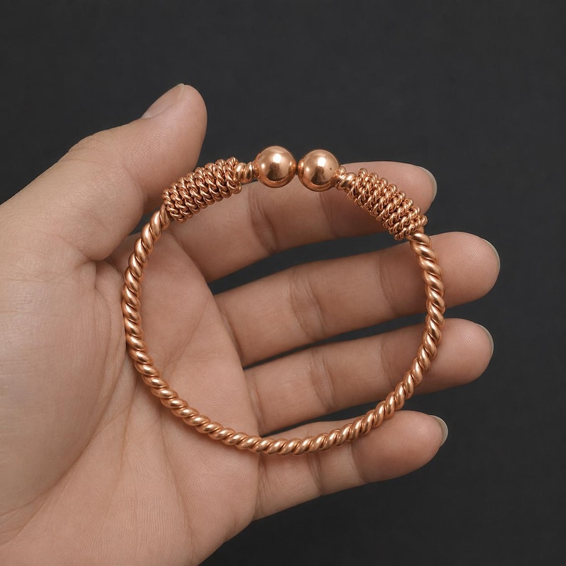 May include: A copper-colored metal cuff bracelet featuring a twisted rope design throughout the band. The open-ended bracelet terminates in two smooth, polished spherical beads, with a coiled wire texture accenting the ends near the spheres.