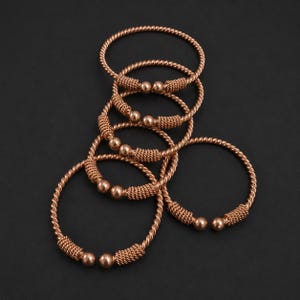 May include: A collection of five copper-toned metal bangles featuring a twisted rope-like texture. Each open-ended bracelet is finished with a smooth, spherical bead at both ends, connected to a section of tightly coiled wire detailing.