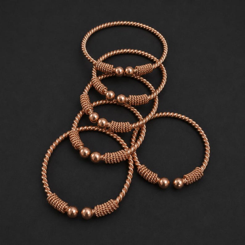 May include: A collection of five copper-toned metal bangles featuring a twisted rope-like texture. Each open-ended bracelet is finished with a smooth, spherical bead at both ends, connected to a section of tightly coiled wire detailing.