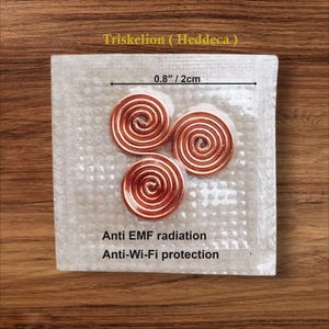 Könnte beinhalten: Drei kupferfarbene Spiralornamente, jeweils etwa 2 cm Durchmesser, liegen auf einer weißen, strukturierten Oberfläche. Der Text lautet "Triskelion (Heddeca)", "Anti EMF radiation" und "Anti-Wi-Fi protection". Der Hintergrund ist Holz.
