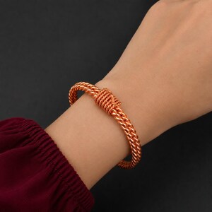 Può includere: Un bracciale color rame caratterizzato da un design a doppio filo intrecciato. I due fili sono uniti al centro da una sezione strettamente avvolta dello stesso filo metallico, creando un aspetto simile a un nodo sul polso.