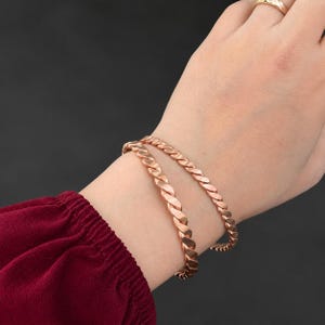 Op de afbeelding: Twee koperkleurige metalen armbanden met een gedraaid, touwachtig ontwerp. De armbanden hebben een gepolijste, metalen afwerking en worden om de pols gedragen. Een eenvoudige, goudkleurige ring is ook zichtbaar aan de vinger van de hand.