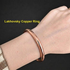 Op de afbeelding: Een gladde, gepolijste koperen armband, aangeduid als Lakhovsky Copper Ring, met een eenvoudig, doorlopend cirkelvormig bandontwerp met een warme, metaalachtige roodbruine afwerking die op de huid rust.