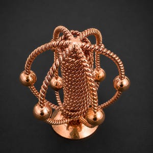 May include: A copper-coloured metal sculpture featuring a central cylindrical pillar wrapped in twisted wire. Several curved, rope-textured arms extend from the top, each ending in a smooth, polished spherical bead, creating an orbital structure.