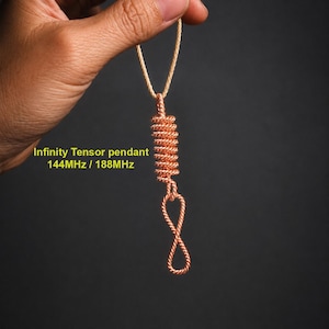Puede incluir: Un colgante de alambre de cobre con una sección cilíndrica enrollada y un bucle en forma de símbolo de infinito en la parte inferior, suspendido por un cordón claro. El texto Infinity Tensor pendant 144MHz / 188MHz aparece impreso en amarillo.