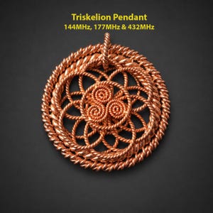 May include: A circular copper-colored pendant featuring a wire-wrapped triskelion design with a twisted rope-style border. The text above reads: Triskelion Pendant, 144MHz, 177MHz & 432MHz. The pendant has a central spiral pattern and a top loop.