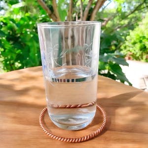 May include: A clear glass with etched floral designs, filled with water, sits on a wooden surface. A copper-colored, twisted metal ring encircles the glass. The background features lush green foliage, suggesting an outdoor setting.