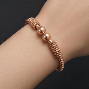 May include: A rose gold-toned bracelet featuring a twisted rope-like texture along the band, accented by two smooth, polished spherical beads positioned centrally. The metal has a warm, metallic sheen and a continuous, flexible design.