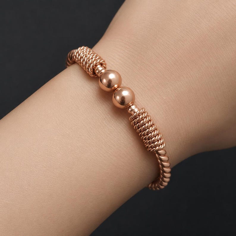 May include: A rose gold-toned bracelet featuring a twisted rope-like texture along the band, accented by two smooth, polished spherical beads positioned centrally. The metal has a warm, metallic sheen and a continuous, flexible design.