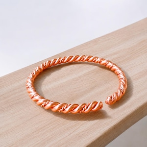 Può includere: Un bracciale a fascia in metallo color rame caratterizzato da un design a corda intrecciata continua. Il bracciale ha una forma circolare aperta con una superficie lucida e riflettente che cattura la luce, evidenziando l'intricata trama a spirale.