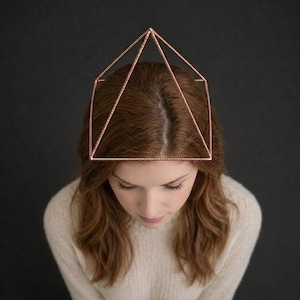 Op de afbeelding: Een geometrisch hoofdaccessoire gemaakt van dunne, roségoudkleurige metaaldraad. De structuur heeft een vierkante basis met vier schuine draden die samenkomen in een centraal punt, waardoor een piramidevorm op het hoofd wordt gevormd.