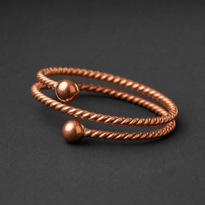 May include: A copper-coloured bypass ring featuring a twisted rope-like texture throughout the band. The ring is open-ended, with a smooth, polished spherical ball at each terminal end, creating a continuous, coiled design with a metallic finish.