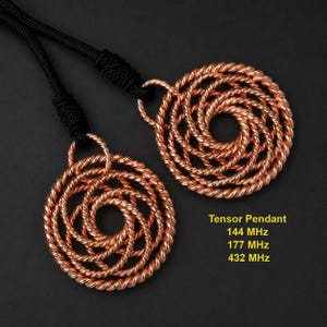 Op de afbeelding: Twee ronde koperdraad hangers met een gedraaid spiraalontwerp, elk bevestigd aan een zwart koord. De tekst op de afbeelding luidt: Tensor Pendant, 144 MHz, 177 MHz, 432 MHz. De hangers zijn gemaakt van meerdere strengen gedraaid koperdraad.