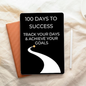 May include: A black tablet with the text "100 Days to Success" and "Track Your Days & Achieve Your Goals" in white. A white stylus and an orange notebook are next to the tablet. The tablet has a white road graphic.