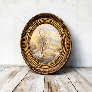 Puede incluir: Pintura enmarcada ovalada que representa un paisaje invernal con un árbol, figuras y un puente. El marco es de color dorado con un diseño ornamentado. La pintura presenta una escena nevada con una paleta de colores apagados.