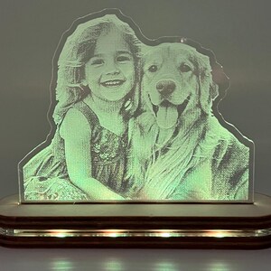 Può includere: Una luce notturna a LED personalizzata con una bambina sorridente e un cane Golden Retriever. L'immagine in acrilico è illuminata da una base in legno con luci integrate. La luce è alimentata da un cavo nero.