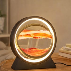 Puede incluir: Una lámpara de arte de arena moderna y circular, con una base y un marco negros. Presenta un globo de vidrio transparente que contiene capas de arena naranja y blanca, iluminado por una luz interna cálida. La lámpara está enchufada.
