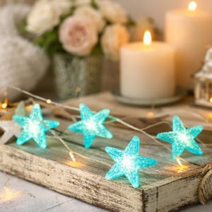 May include: A string of turquoise star-shaped fairy lights on a rustic wooden tray. The lights are illuminated, creating a soft glow. The background includes lit candles, flowers, and a decorative house, suggesting a cozy, festive atmosphere.