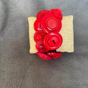 May include: A bracelet constructed from a collection of various red buttons of different sizes and textures. The buttons are layered and connected to form a circular band, featuring smooth, ridged, and floral-shaped designs in a vibrant red hue.