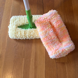 Puede incluir: Una mopa con un mango blanco y verde y dos cabezales rectangulares. Un cabezal es amarillo y el otro naranja, rosa y blanco. La mopa está diseñada para limpiar suelos.