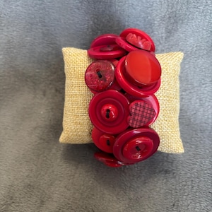 May include: A red button bracelet with various sizes and styles of buttons. The bracelet is displayed on a small, square, tan pillow. The buttons are a mix of glossy and matte finishes.