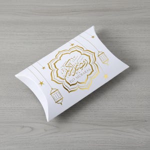 Könnte beinhalten: Weiße Kissenbox mit goldfarbenen Akzenten. Die Box zeigt den Schriftzug "Eid Mubarak" in Gold, zusammen mit Stern- und Laternenmotiven. Ideal zum Verschenken.