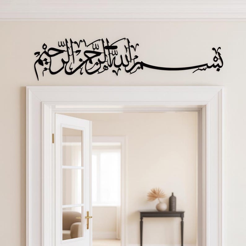 May include: Black Arabic calligraphy wall art above a white doorway. The script is a flowing design, likely a religious phrase. The doorway is open, revealing a glimpse of a room with neutral-toned walls and decor.