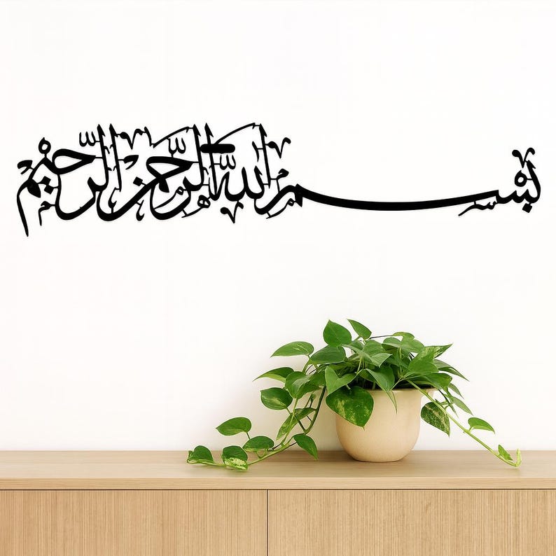 May include: Black Arabic calligraphy wall art. The script is a flowing design, mounted on a white wall above a wooden cabinet. A potted plant with green leaves is in the foreground. The calligraphy is a decorative piece.
