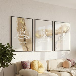 Puede incluir: Tres impresiones artísticas enmarcadas con caligrafía árabe blanca sobre un fondo abstracto beige y dorado. Las impresiones se exhiben sobre un sofá beige con cojines decorativos y una planta.