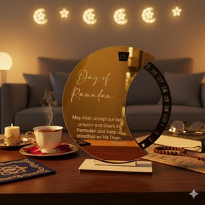 May include: A gold crescent moon-shaped Ramadan calendar with the text "Day of Ramadan" and a prayer. The calendar displays the date and has a rotating dial. A cup of tea, candle, and decorative lights are also visible.