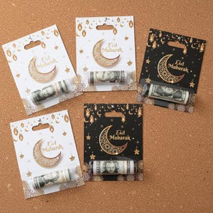 Könnte beinhalten: Eid Mubarak Geschenkkarten mit einer aufgerollten Banknote. Die Karten zeigen ein Mond-Design und den Schriftzug "Eid Mubarak". Die Karten sind weiß und schwarz mit goldenen Akzenten.
