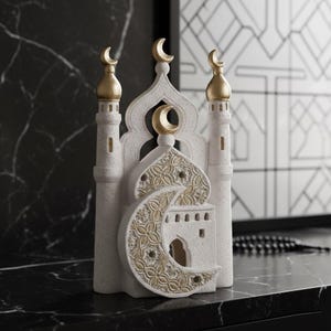 Puede incluir: Una figura decorativa de mezquita blanca y dorada. La figura presenta tres minaretes rematados con medias lunas doradas. La estructura central tiene una media luna y patrones dorados intrincados. La base es blanca con un acabado texturizado.