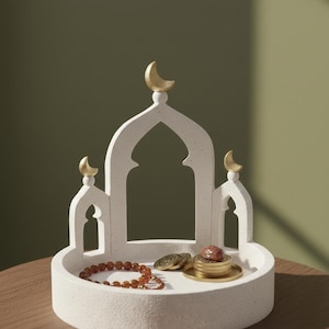 Op de afbeelding: Een witte, getextureerde decoratieve tray met een moskee-achtig ontwerp, bekroond met gouden halve manen. De tray bevat een kralenarmband, munten en een klein, opgestapeld item. De achtergrond is een gedempt groen.
