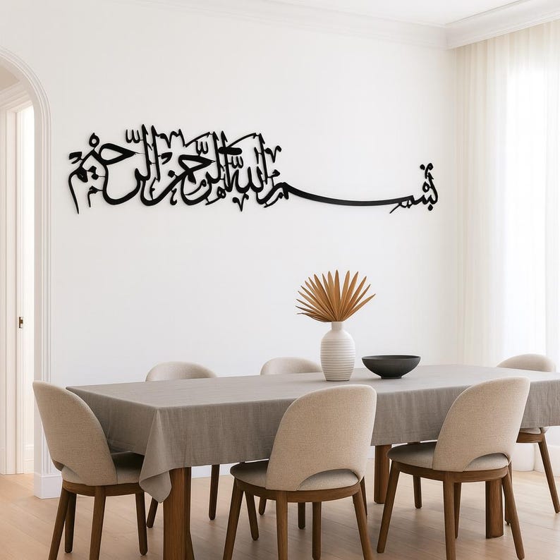 May include: Black metal wall art featuring Arabic calligraphy script, mounted on a white wall. The artwork is above a dining table set with a gray tablecloth, beige chairs, and a decorative vase.