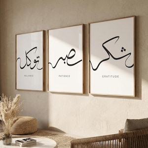 Op de afbeelding: Een set van drie verticale wandposters in lichte houten lijsten. Elke witte print bevat zwarte Arabische kalligrafie met de bijbehorende Engelse woorden Reliance, Patience en Gratitude in kleine zwarte letters onder elk schrift.