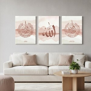 May include: Three framed canvas art pieces with Arabic calligraphy in shades of brown and white. The artwork features intricate script and abstract watercolor backgrounds. The pieces are displayed above a white sofa in a living room setting.