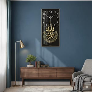 Op de afbeelding: Een rechthoekige zwarte wandklok met gouden Arabische kalligrafie en witte cijfers. De klok heeft een gouden lijst en gouden wijzers. De klok hangt aan een blauwe muur.