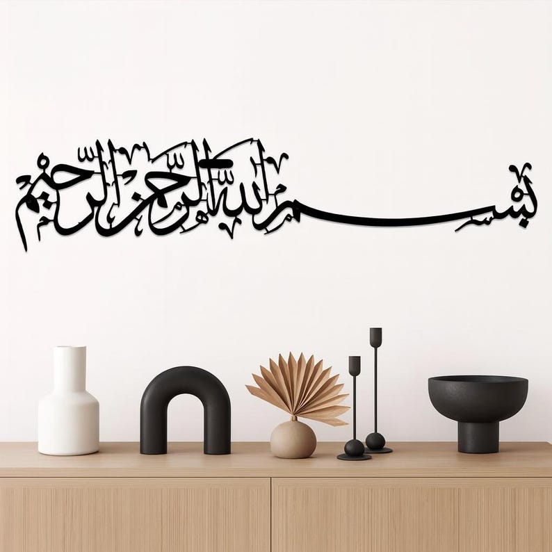 May include: Black metal wall art featuring Arabic calligraphy script. The script is mounted on a white wall above a wooden cabinet. Decorative items on the cabinet include a white vase, a black arch, a beige vase with a fan-shaped decoration, and black candle holders.