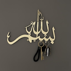 May include: A gold-coloured metal key holder in the shape of Arabic calligraphy. The key holder is mounted on a grey wall and holds several keys. The calligraphy reads "Bismillah" in a stylized script.