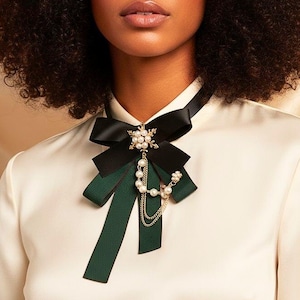 Puede incluir: Una pajarita negra y verde con detalles de perlas y cadena dorada. La pajarita se lleva sobre una blusa color crema. La pajarita tiene un lazo negro en la parte superior y un lazo verde debajo. Un accesorio de moda.