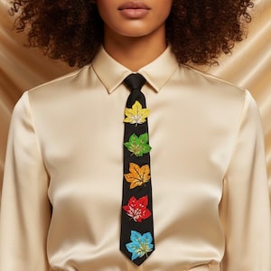 May include: A black tie adorned with embroidered colorful leaf designs. The leaves are yellow, green, orange, red, and blue. The tie is worn with a cream-colored button-down shirt.